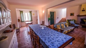 Zona pranzo con un tavolo lungo, sedie e tovaglia decorata. C'è un angolo cottura con piano cottura, lavello e pensili. Una scala conduce a un piano superiore.