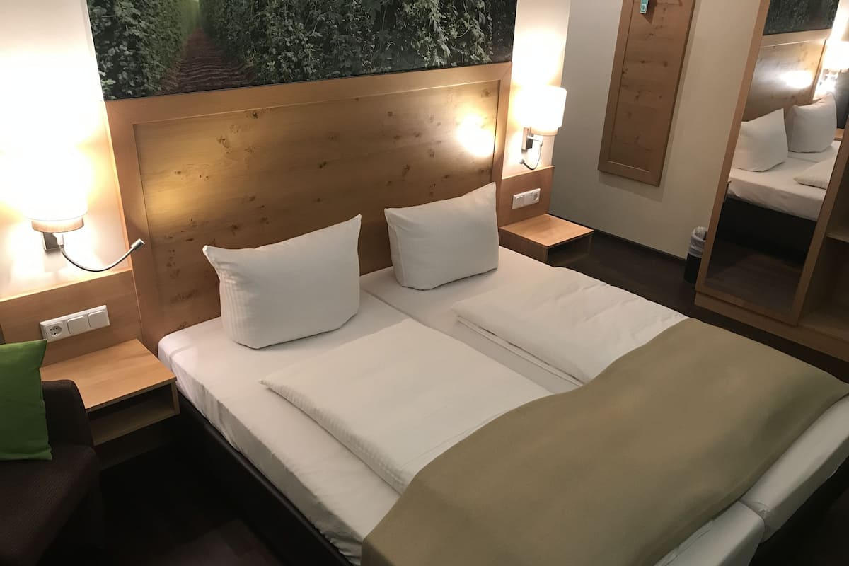 Ein ordentlich bezogenes Doppelbett mit weißen Bettwäsche, ein Holz-Kopfteil, Nachttischlampen und ein kleiner Nachttisch mit einem grünen Kissen.