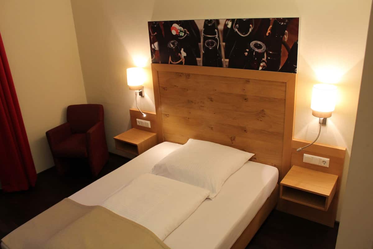 Ein Hotelzimmer mit hölzernem Kopfteil, einem Einzelbett mit weißen Bettwäsche, zwei Nachttischlampen, einem roten Sessel und einem Wandbild.