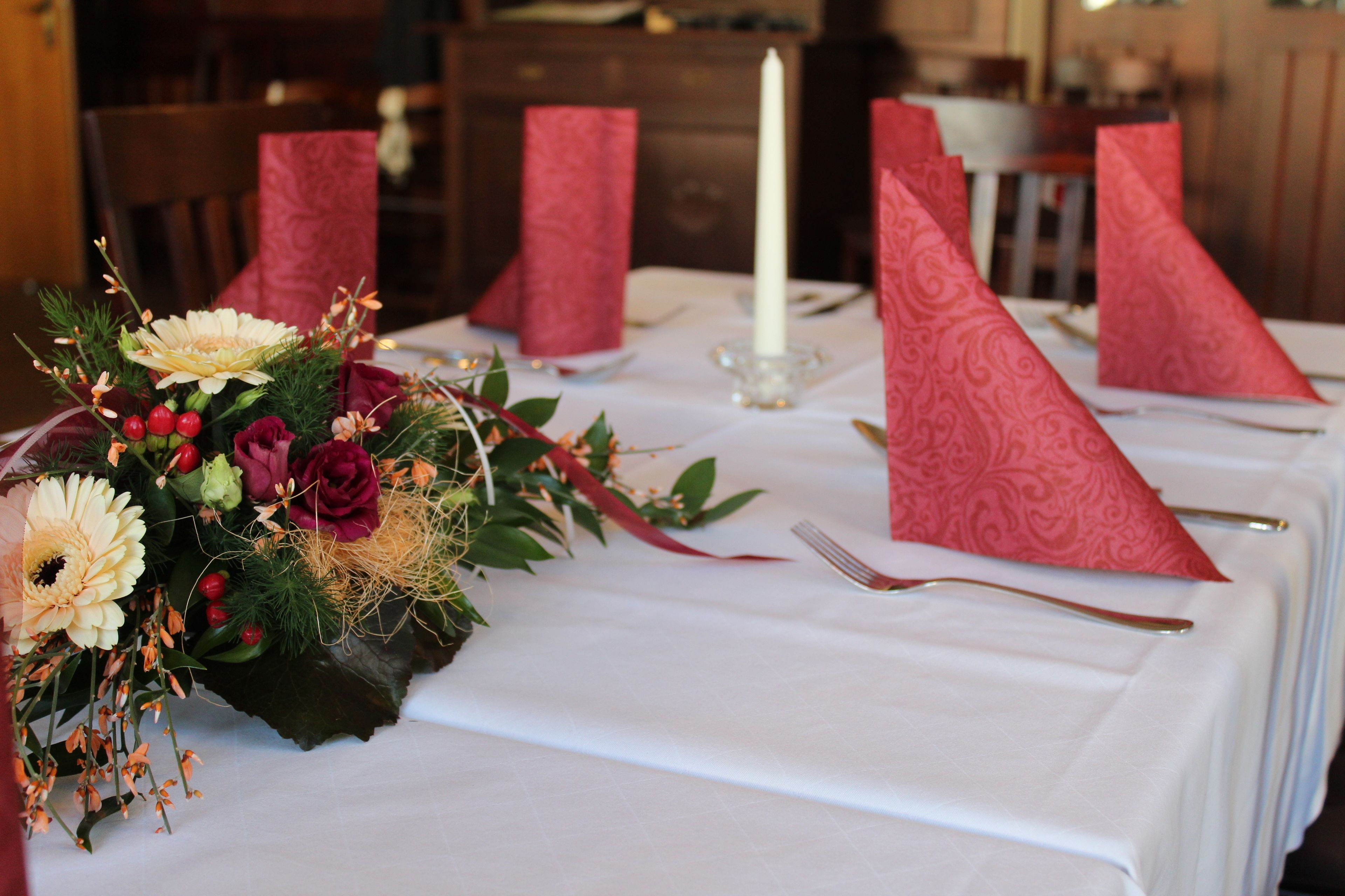 Een gedekte tafel met een witte tafelkleed, rode servetten, een kaars en een bloemstuk.