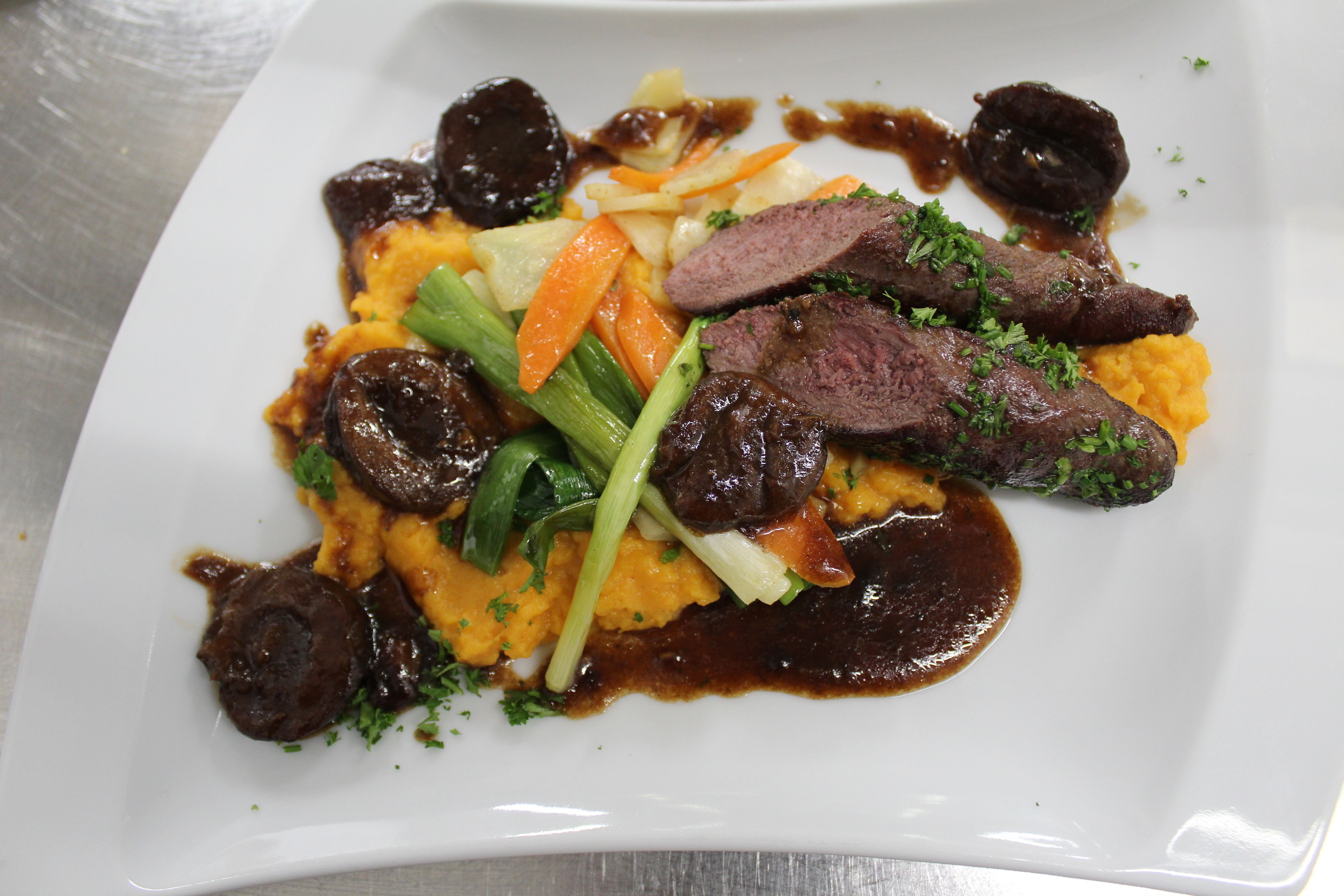 A square white plate with a dish consisting of sliced meat, vegetables, and a sauce.