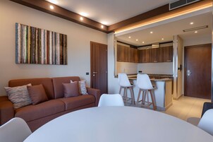 Una sala moderna con un sofá marrón, una mesa redonda y sillas blancas. Se ve un área de cocina con armarios de madera y una encimera a través de una puerta abierta.