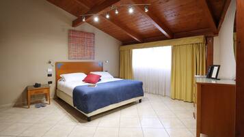 Quarto com teto de madeira, cama com colcha azul, mesa de cabeceira com telefone, criado-mudo e janela com cortinas.