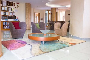 Un hall con una mesa de centro redonda de vidrio, varios sillones, una estantería con diplomas enmarcados y una alfombra colorida.