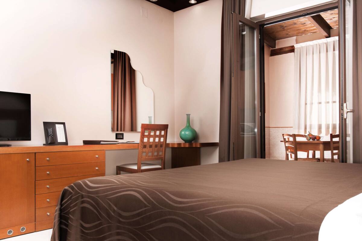 Ein Hotelzimmer mit einem Bett, einem Schreibtisch mit Computer, einem Fernseher und einem Fenster mit Vorhängen.