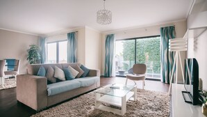 Ein modernes Wohnzimmer mit einem beigen Sofa, einem Glastisch und einem großen Fenster mit Vorhängen.