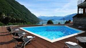 Ein rechteckiger Swimmingpool mit klarem, blauem Wasser, umgeben von einer Holzterrasse und Liegestühlen, vor einer Berglandschaft und einem See.