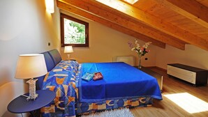 Ein Schlafzimmer mit schräg gedachter Decke, einem Bett mit blauer Bettdecke, einem Nachttisch mit Lampe und einem Fenster mit Blick ins Grüne.