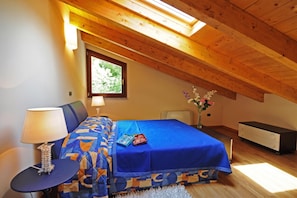 A bedroom with a sloped ceiling, a bed with a blue cover, a bedside table with a lamp, and a window with a view of greenery. - Residence Eden Camelia 7, Argegno, Italy (Argegno)