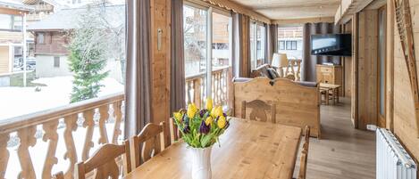 A wooden dining table with a vase of flowers, wooden chairs, a TV, and a view of the outside.