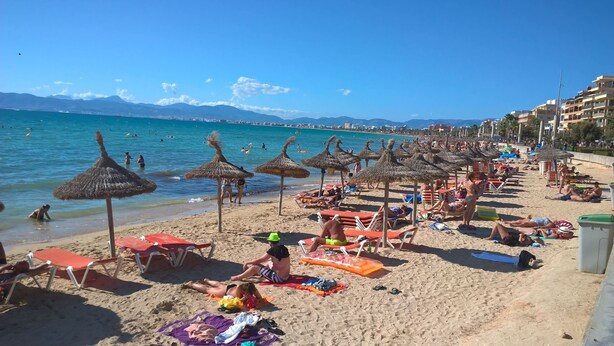 Une plage bordée de rangées de chaises longues et d’ombrelles en chaume, une mer d’un bleu limpide et des montagnes à l’horizon.