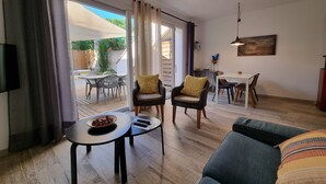 Un salon comprenant un canapé, une table basse ronde, deux fauteuils, une table à manger avec des chaises, et un coin cuisine visible à travers une porte coulissante ouverte.