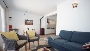 Un salon avec des fauteuils en osier, un canapé bleu, une table basse agrémentée d’objets décoratifs, et une cuisine en arrière-plan.