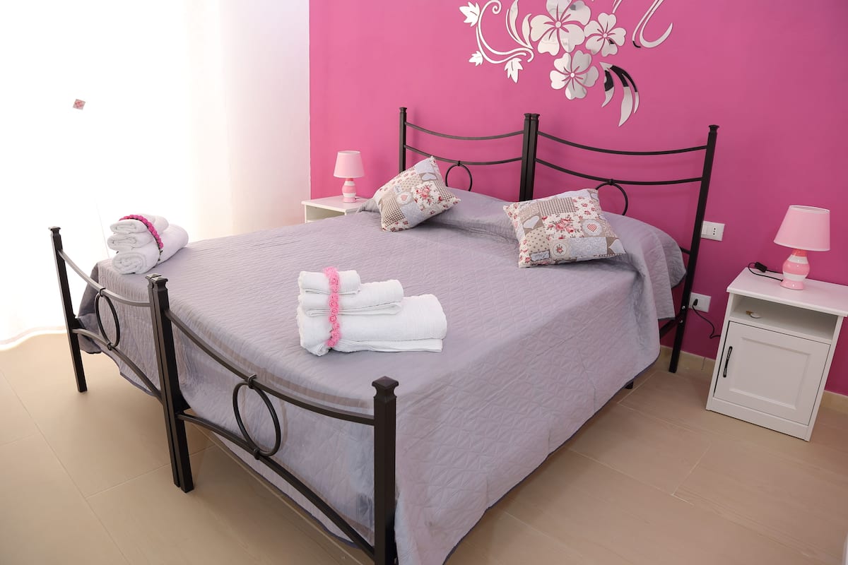 Un lit bien fait, avec des oreillers et des serviettes pliées, une table de chevet avec une lampe et un mur orné d’un motif floral.