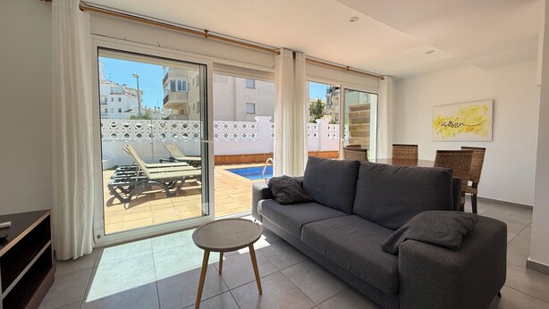 A modern living room with a grey sofa, a small round table, and a view of a pool and buildings outside. - Agi Las Acacias 2401 Apartamento for rent in Roses Girona. (Rosas / Roses)
