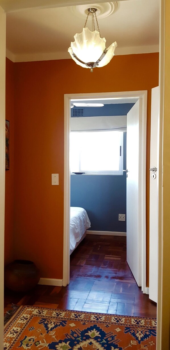 Um corredor com um tapete estampado, uma cama no quarto vizinho e um lustre no teto.