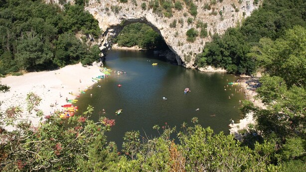 A natural arch over a river with boats and people on the shore.