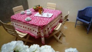 A table with a patterned tablecloth, surrounded by chairs, in a room with a plain wall and a brown floor.