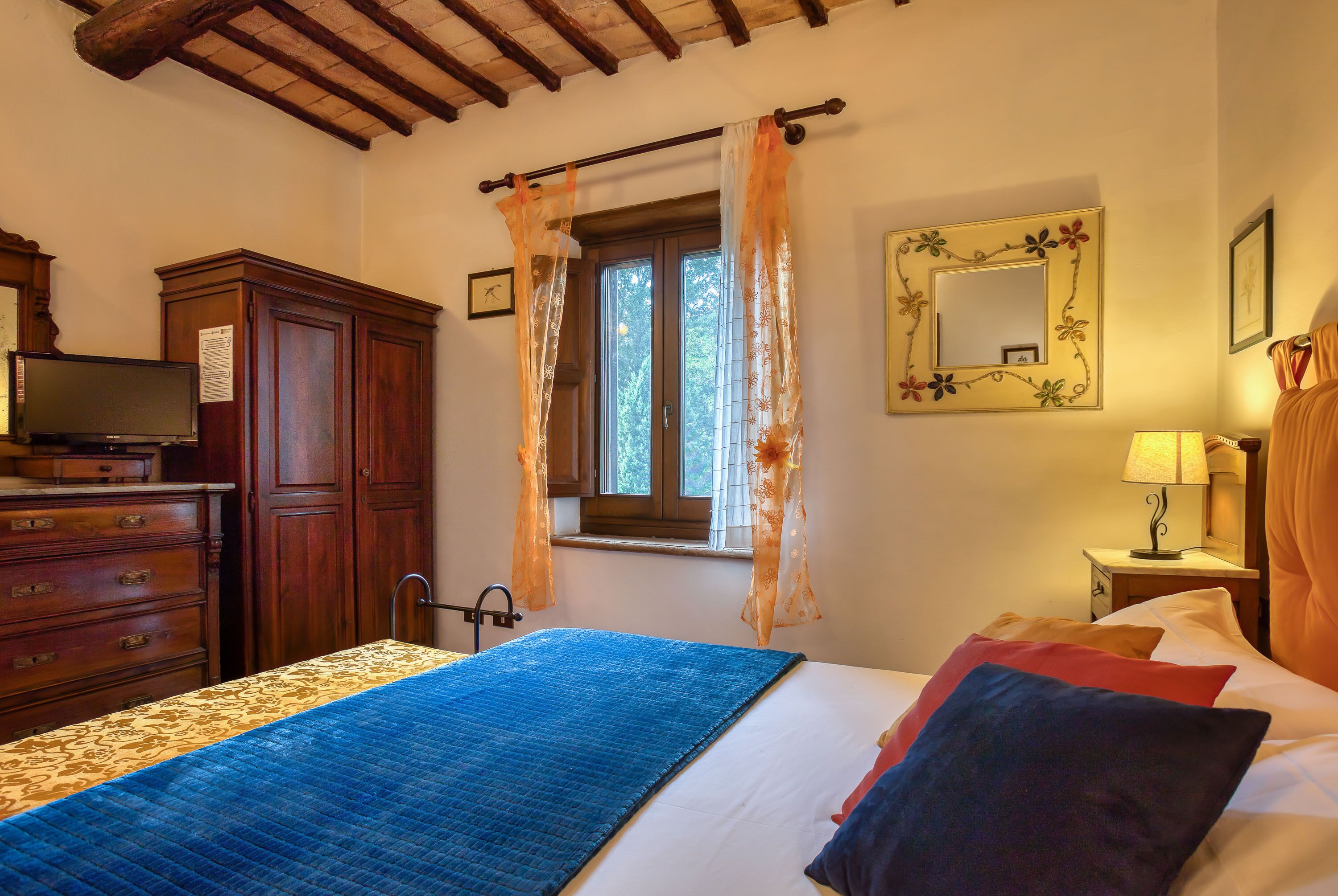 Ein Schlafzimmer mit Bett, Holzschrank, Fernseher auf einer Kommode, Fenster mit Vorhängen, Nachttischlampe und Spiegel an der Wand.