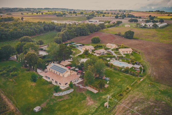 Vista aérea de una extensa propiedad con varias construcciones, incluyendo una casa principal con techo de paneles solares, una piscina y jardines circundantes.