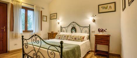 Un dormitorio con cama, piso de madera, cortinas, mesita de noche, lámpara de pared e imágenes enmarcadas.