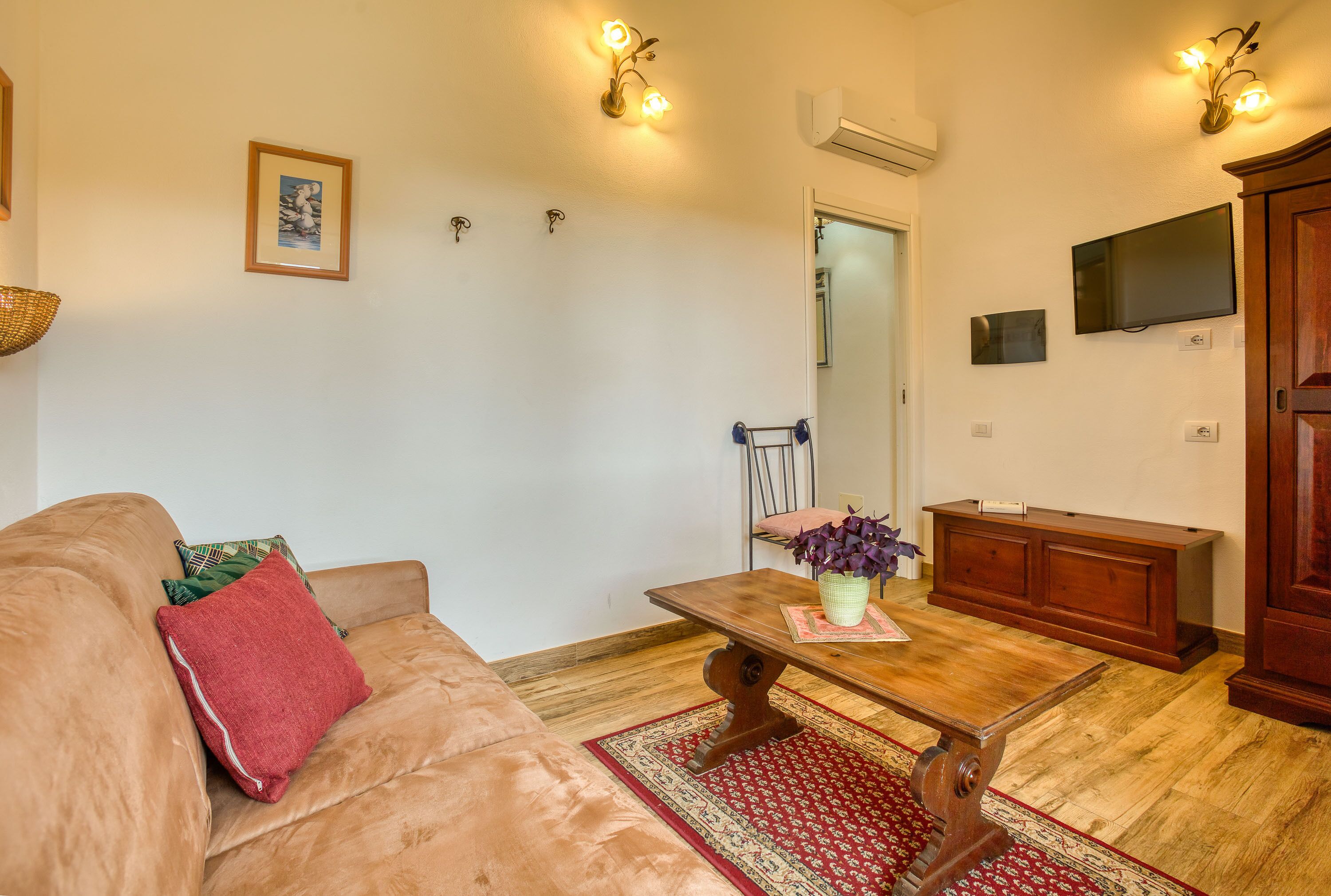 A living room with a brown sofa, a wooden coffee table with a vase, a red rug, a wooden cabinet, and a TV mounted on the wall.