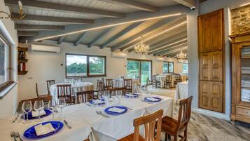 A dining area with tables set for a meal, wooden beams on the ceiling, and large windows with a view of greenery.