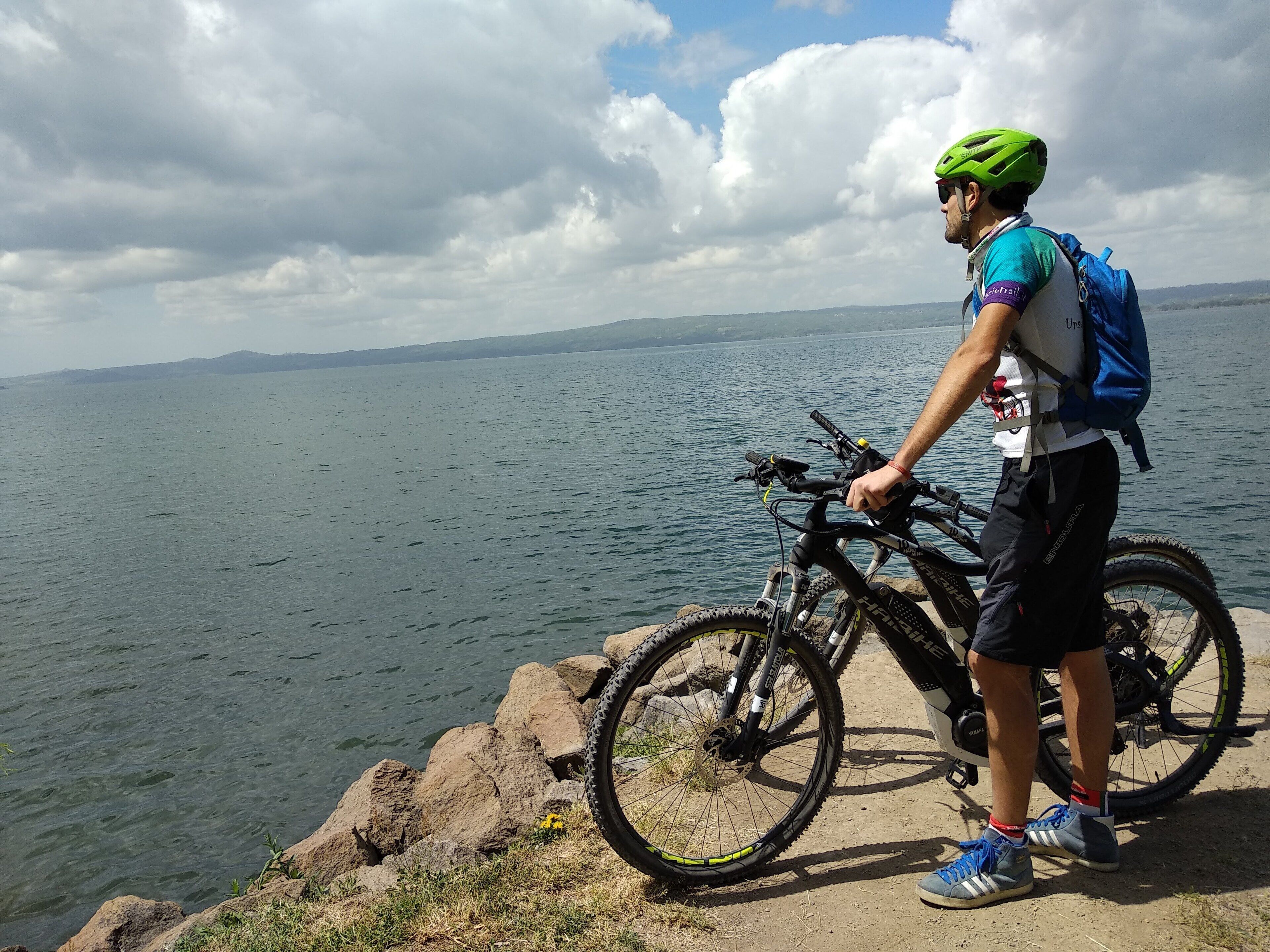 A person standing next to a mountain bike on a rocky shore with a lake and distant mountains in the background.