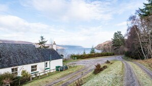 A rural house with a white exterior and dark roof, a gravel driveway, and a view of a lake and hills in the background. - Lovely foresters cottage, amazing Loch Ness views, private beach, pets welcome (Inverfarigaig,   Loch Ness)