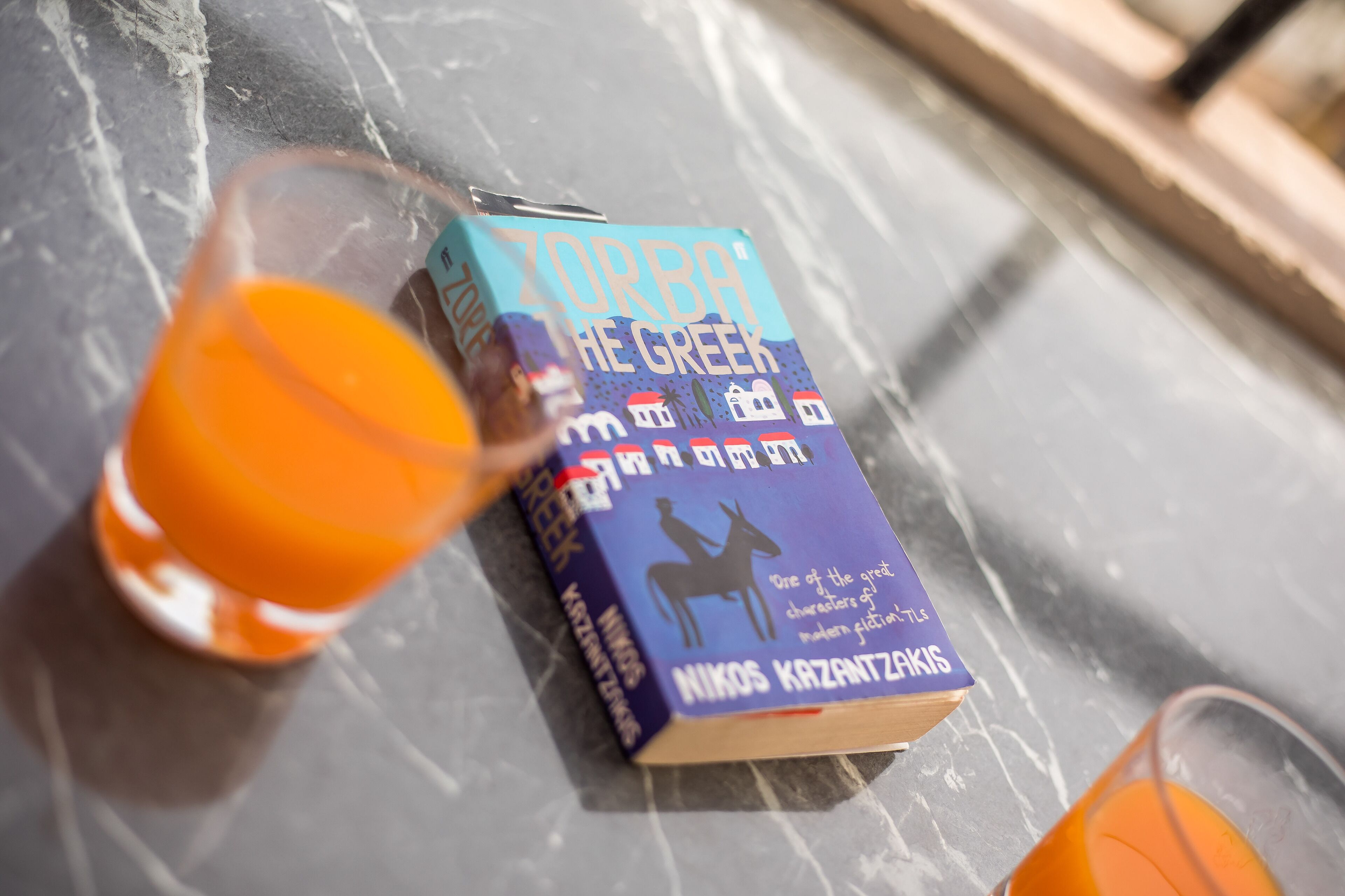 Un libro titulado 'Zorba el Griego' de Nikos Kazantzakis sobre una mesa de mármol, junto a un vaso de jugo de naranja.