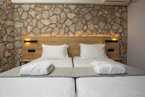 Cama doble con sábanas blancas y dos almohadas, pared de piedra al fondo y cabecera de madera con dos mesitas de noche.