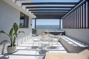 Un patio al aire libre iluminado por el sol, con una mesa, sillas, una planta en maceta y vistas al mar.