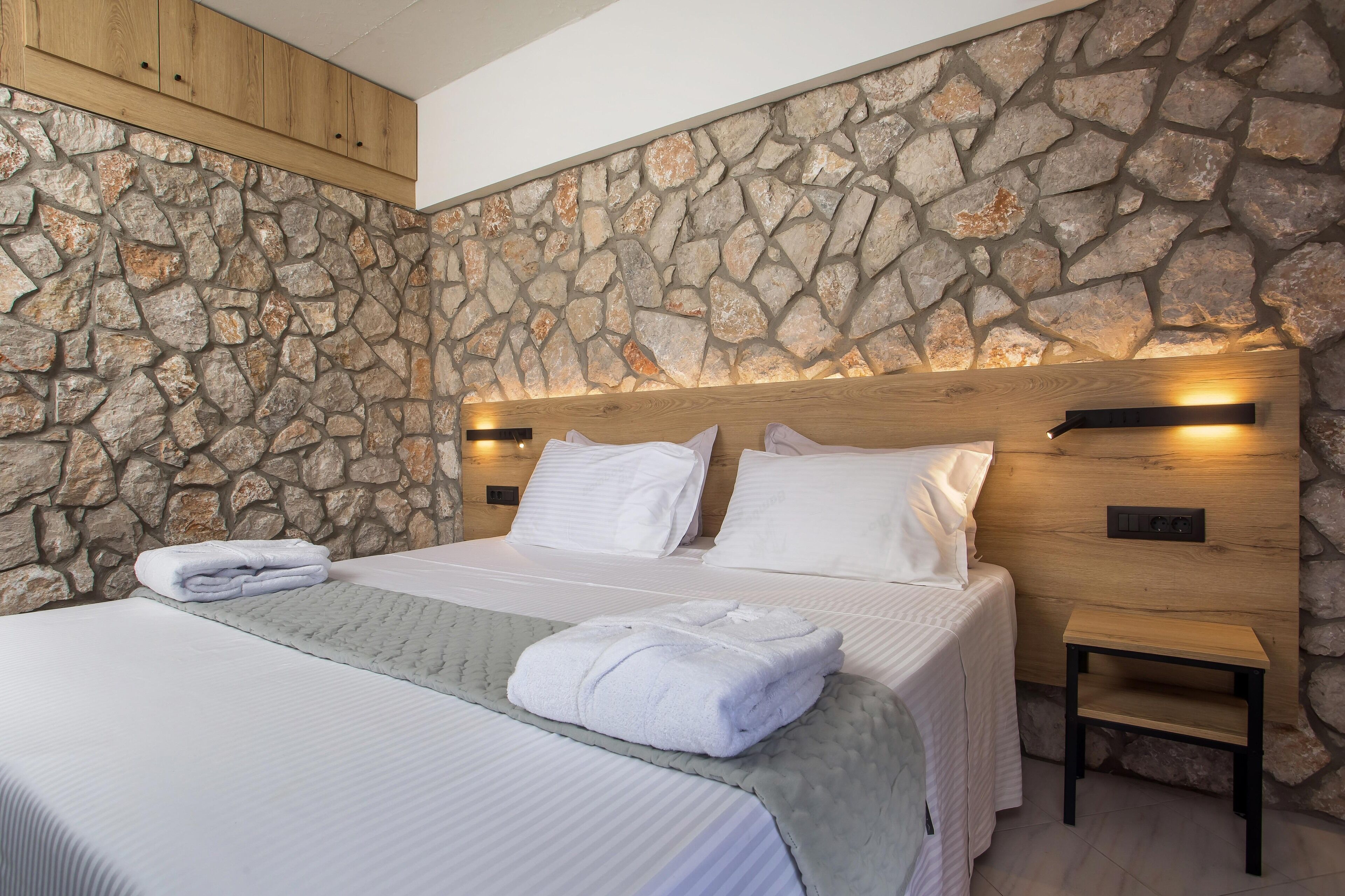 A bedroom with a stone wall, wooden headboard, two beds, white bedding, and a small bedside table.