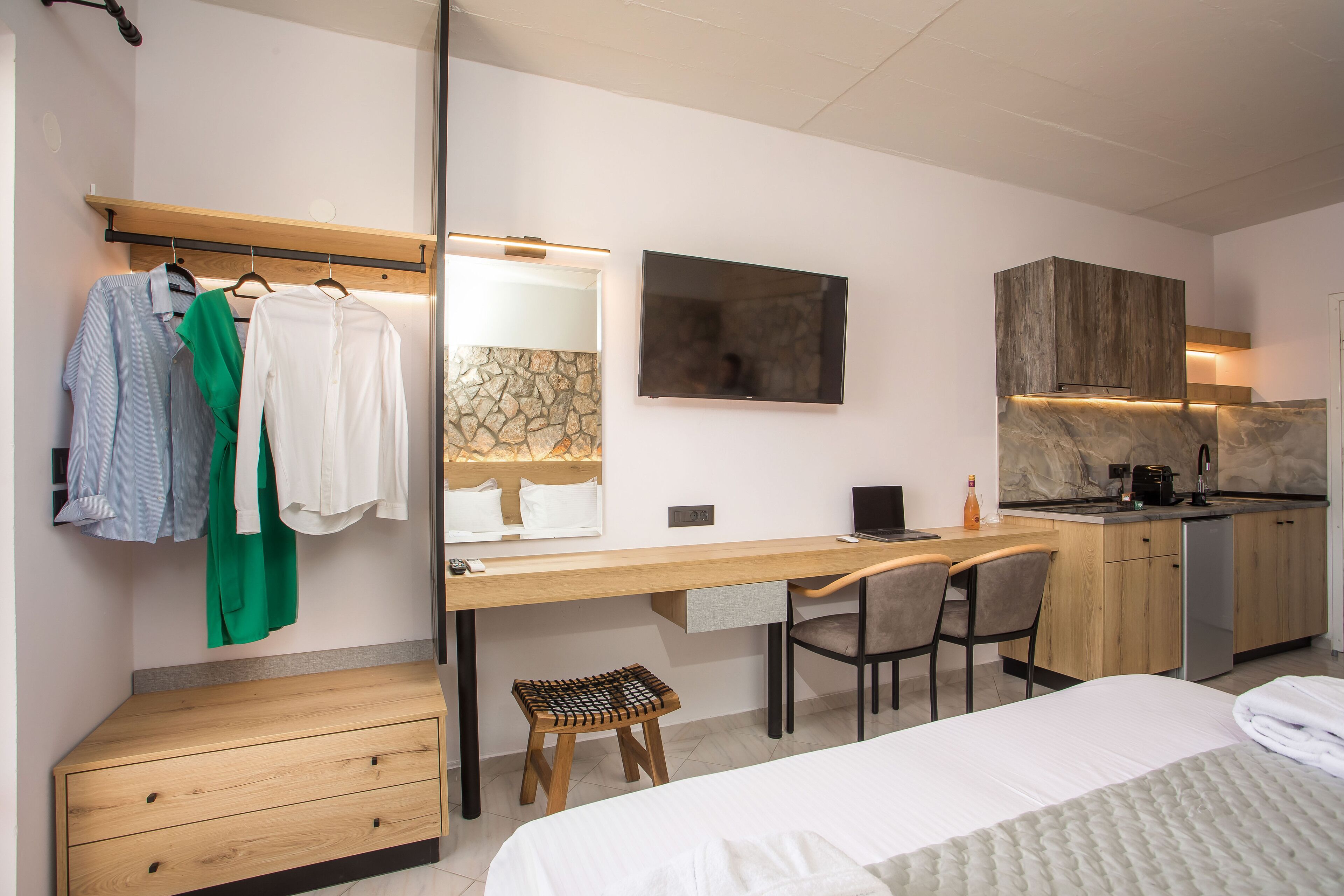 A modern hotel room with a bed, a desk with a chair, a small stool, a wardrobe, a mirror, and a kitchen area with a countertop and cabinets.