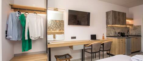 A modern hotel room with a bed, a desk with a chair, a small stool, a wardrobe, a mirror, and a kitchen area with a countertop and cabinets.