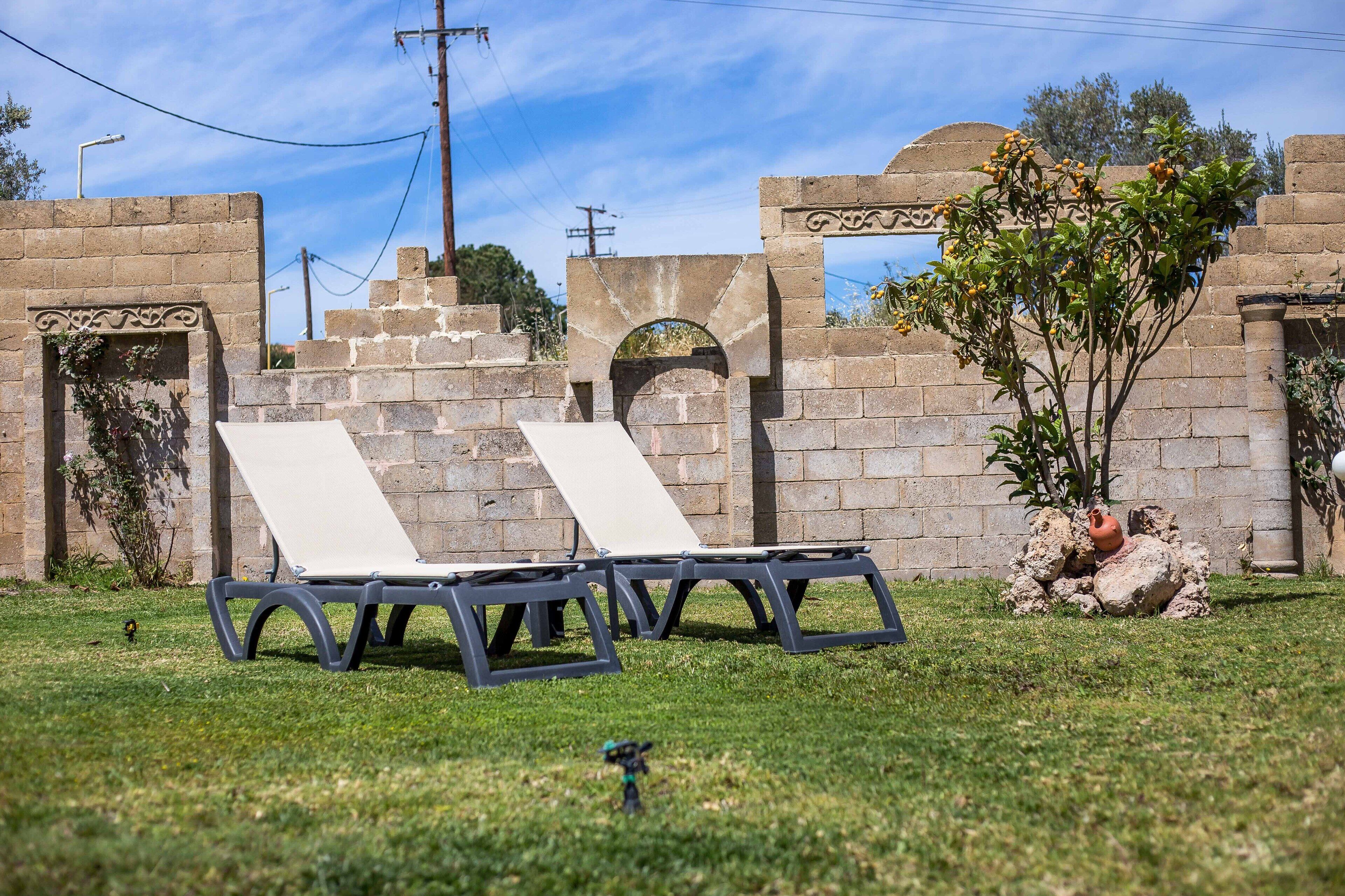 Deux fauteuils de jardin sont placés dans un espace verdoyant, avec un mur de pierre et un arbre aux fleurs jaunes en arrière-plan.