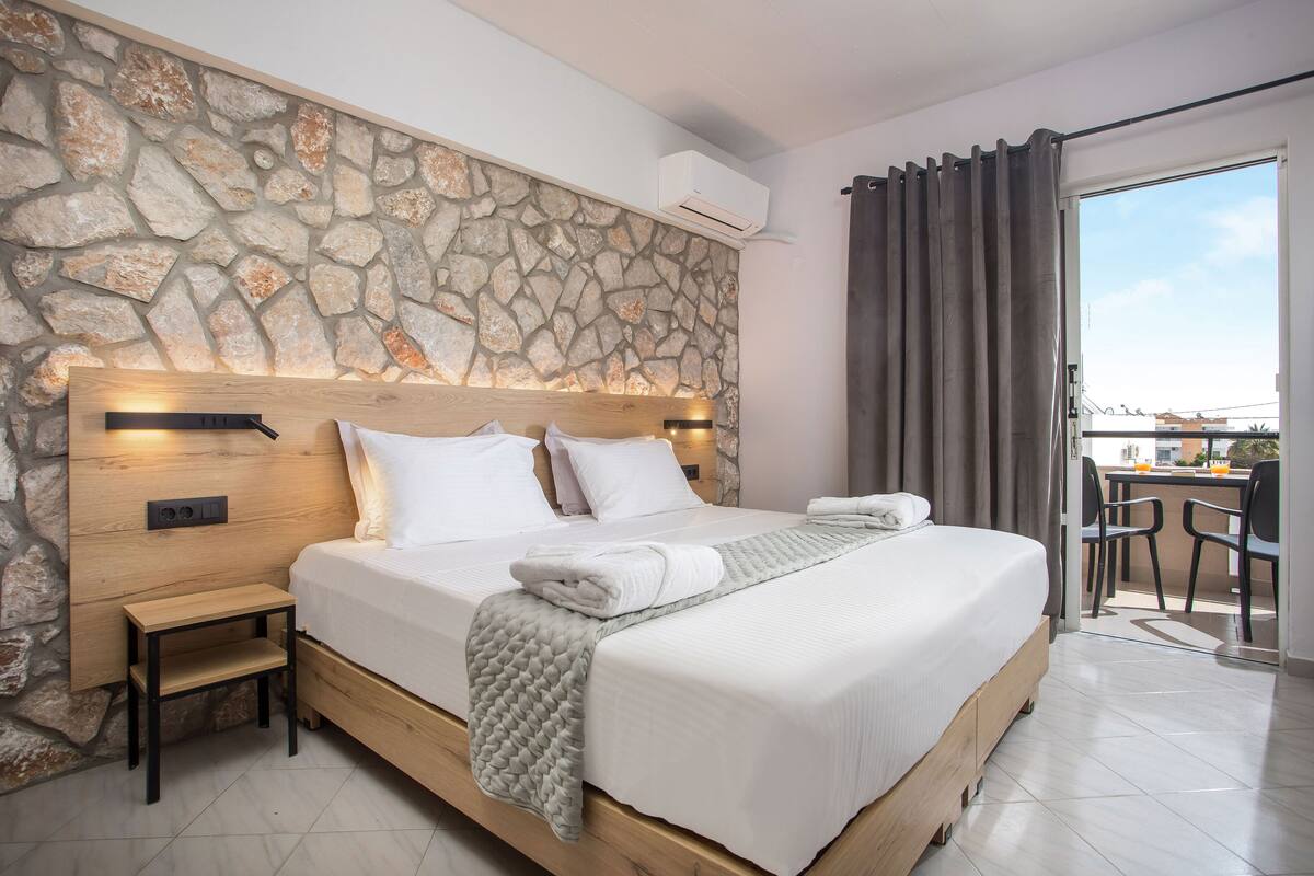 A modern bedroom with a stone wall, a wooden bed frame, a bedside table, and a view of the balcony.