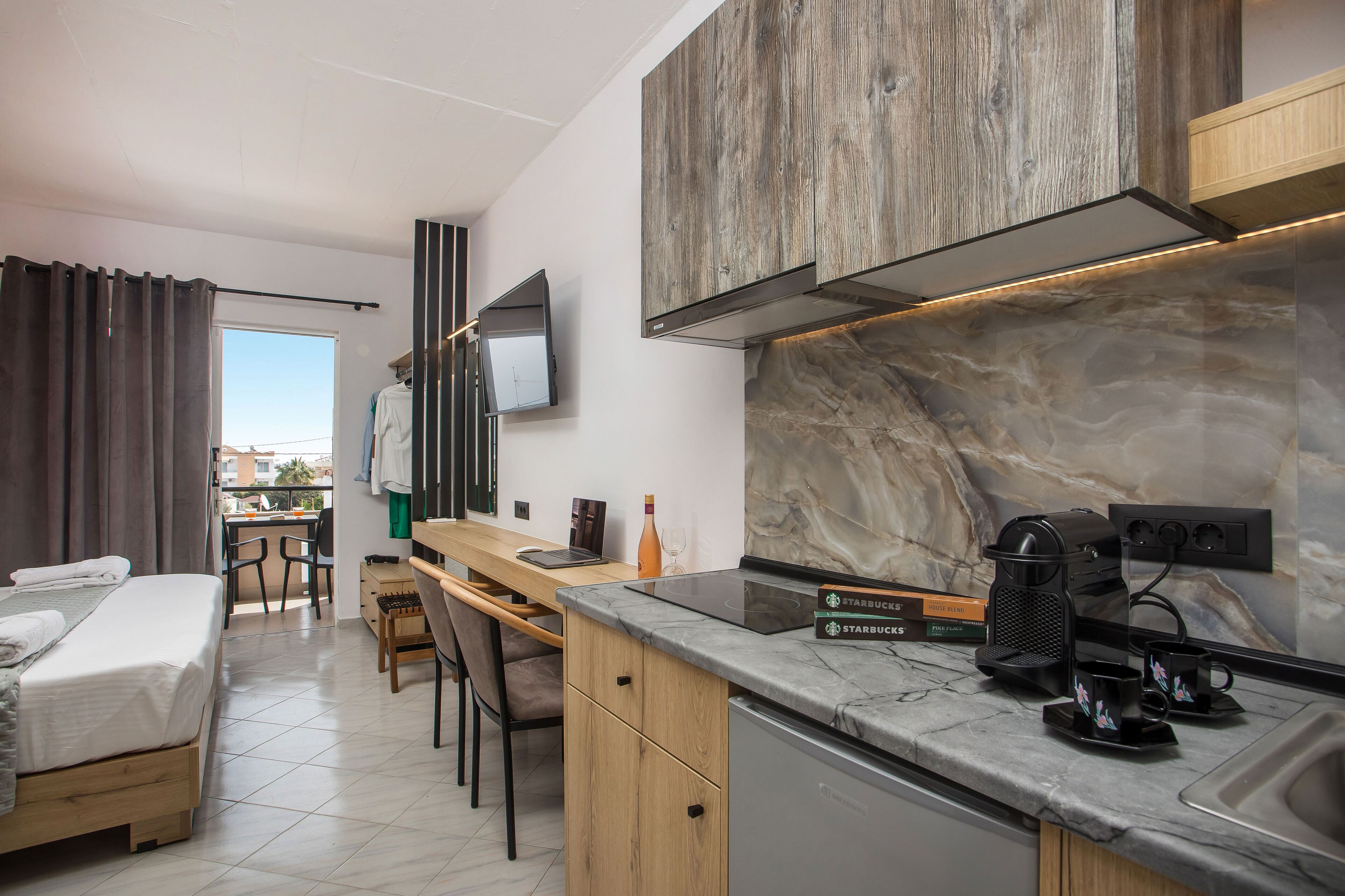 A modern hotel room with a bed, a desk, a chair, a kitchen area with a coffee maker, and a TV mounted on the wall.
