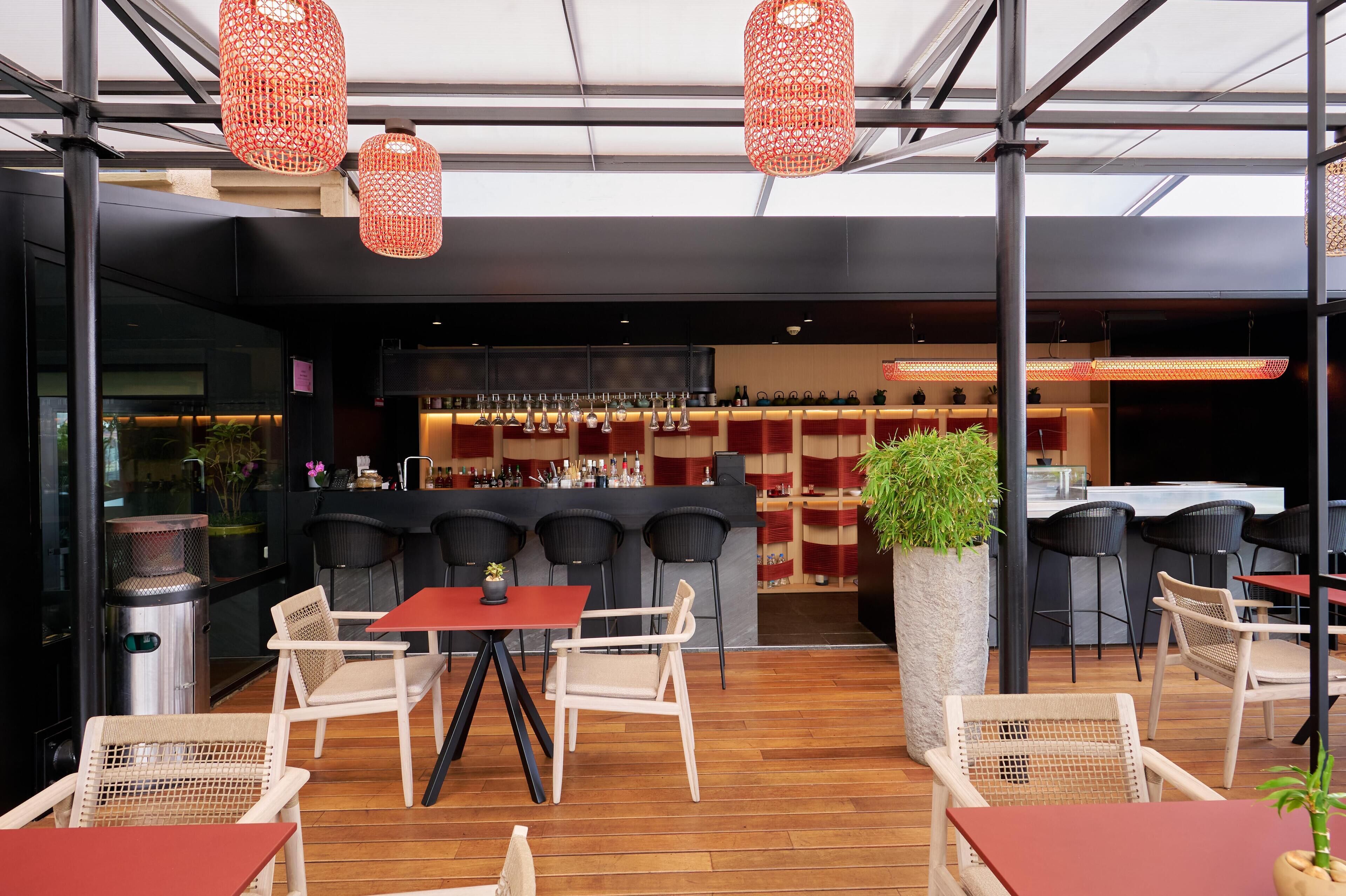 Un espace extérieur aménagé avec des chaises et des tables en osier, un bar avec des tabourets et des lanternes rouges suspendues.