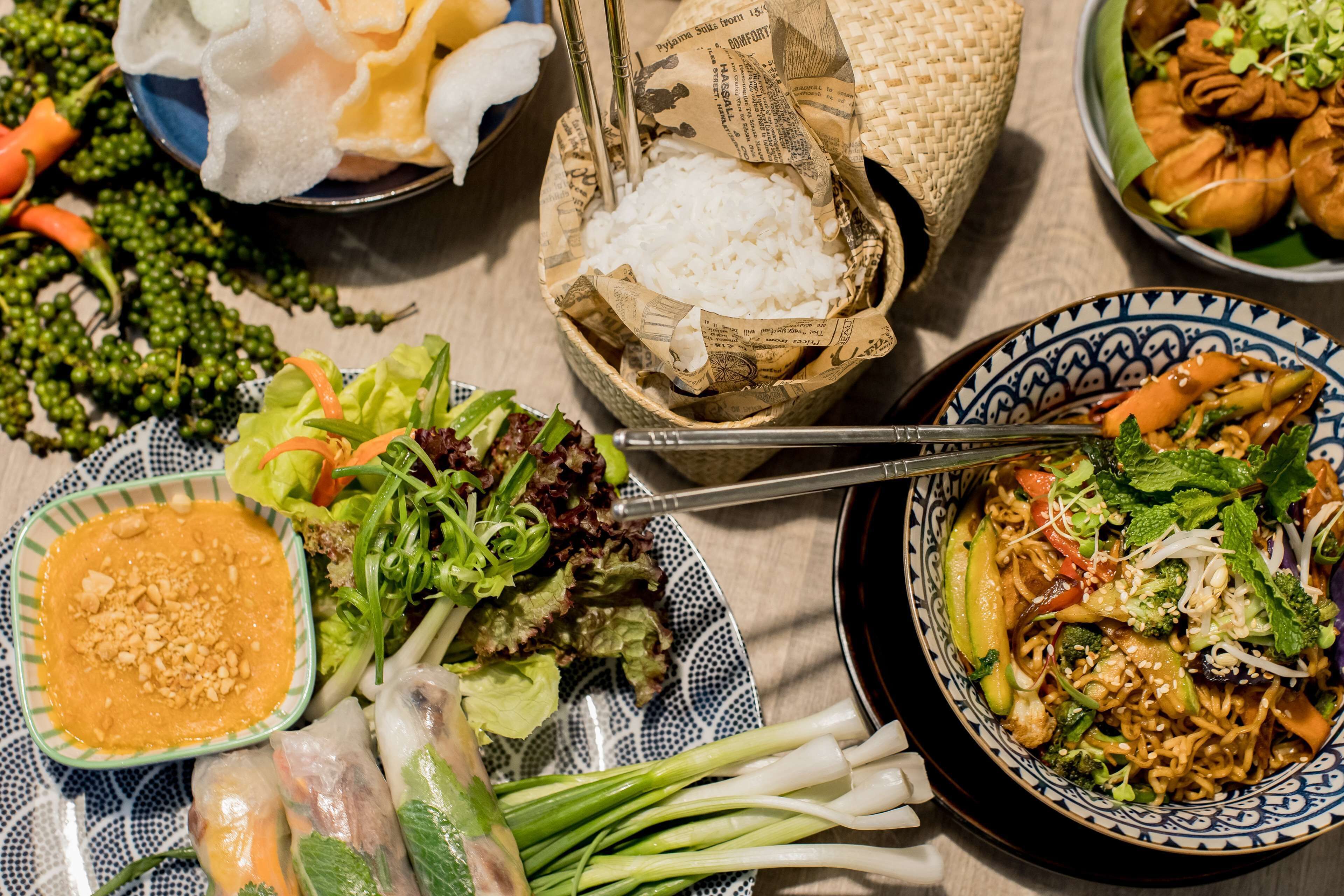 A table with various dishes including rice, noodles, vegetables, and spring rolls.