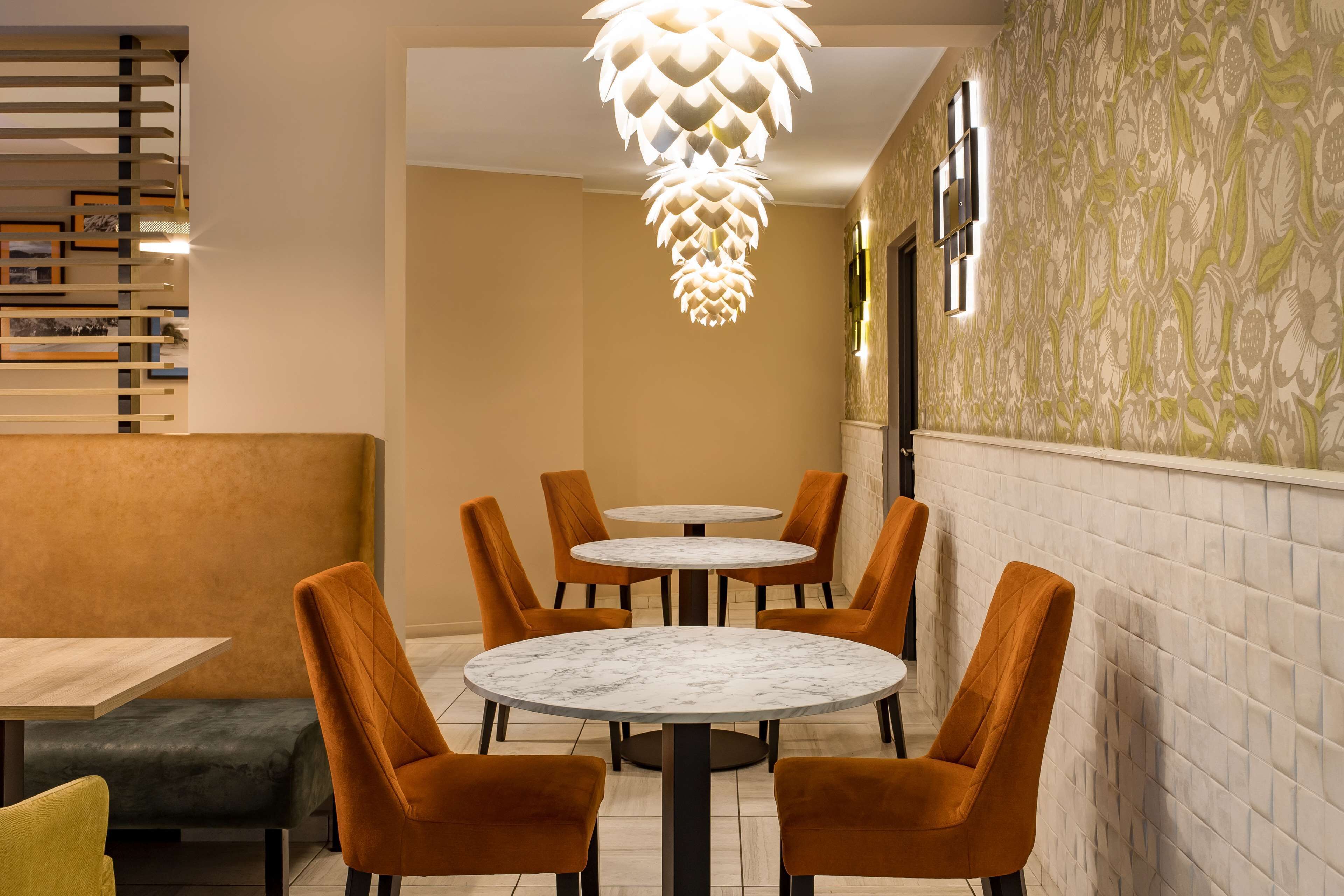A dining area with round tables and orange chairs, a patterned wall, and a unique ceiling light fixture.