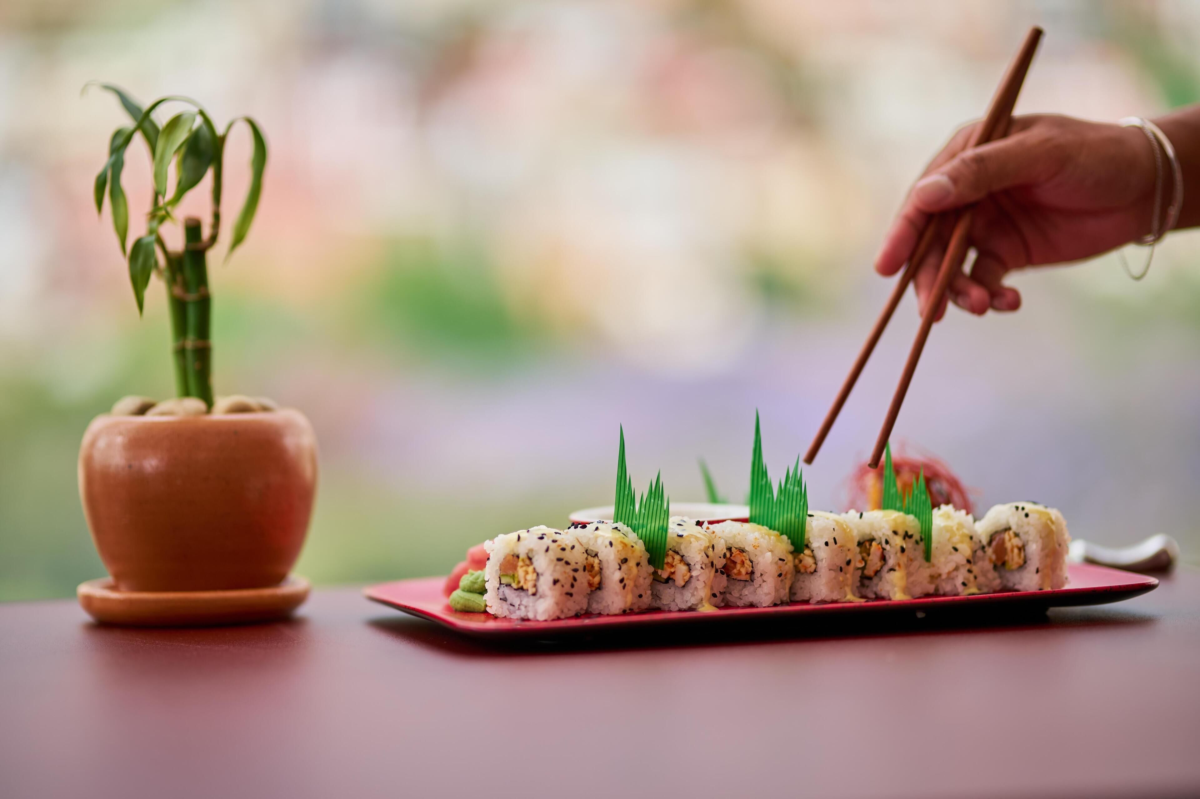 Egy sushi-tál étkészletként, pálcikákkal, és egy virágos növény egy cserépben, mindez egy asztalon.