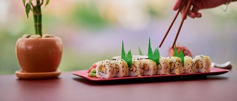 Egy sushi-tál étkészletként, pálcikákkal, és egy virágos növény egy cserépben, mindez egy asztalon.