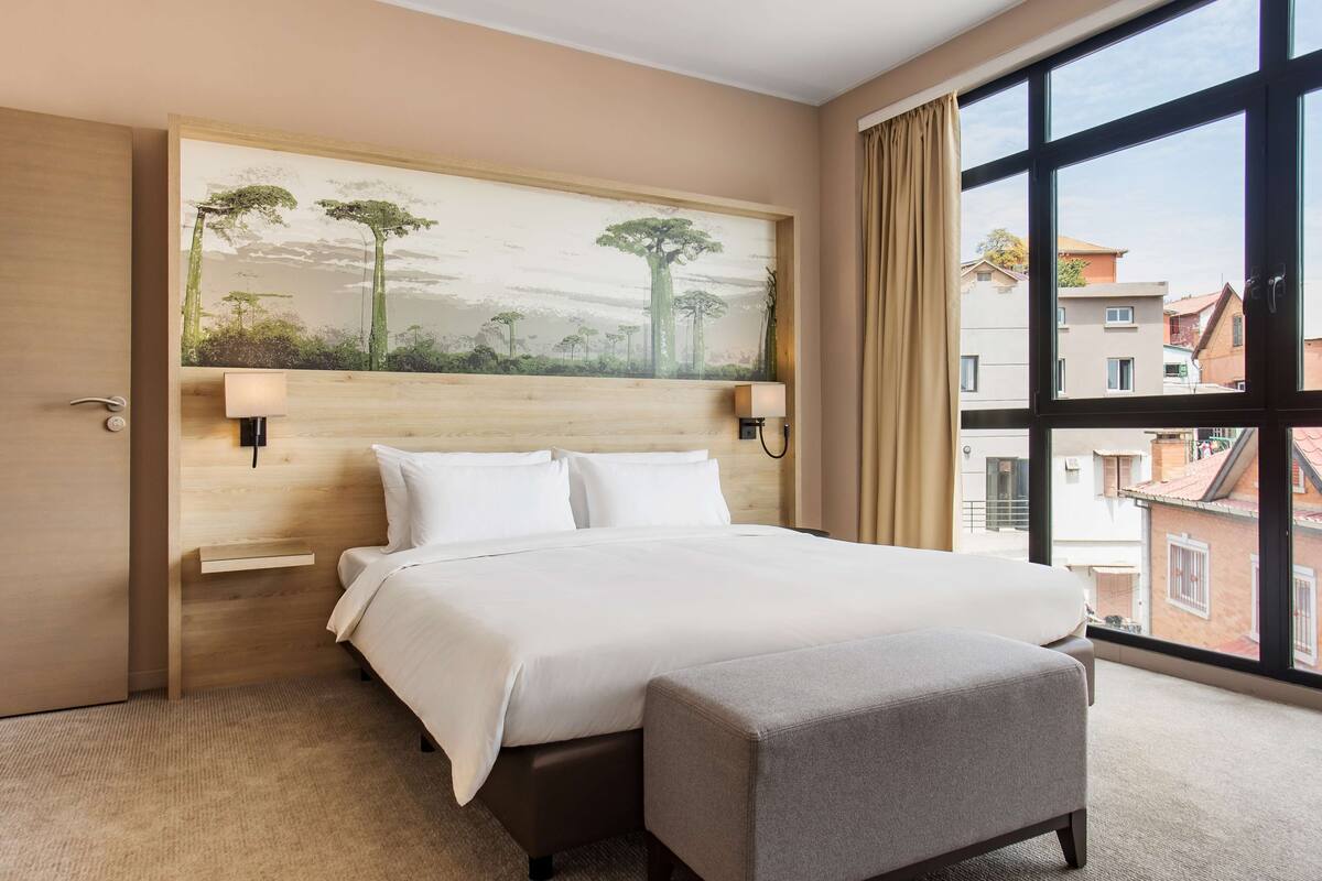 Une chambre d’hôtel avec un grand lit, une table de chevet, une commode, une lampe fixée au mur et une grande fenêtre donnant sur des immeubles.