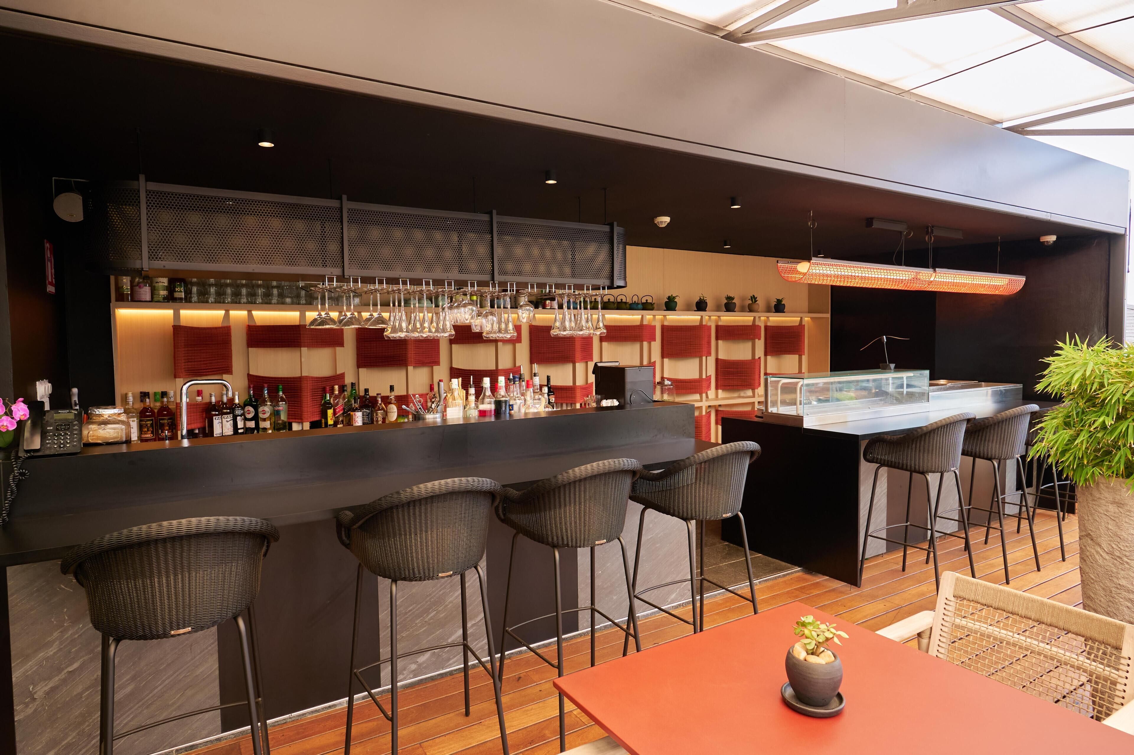 Un espace bar moderne avec un comptoir noir, des tabourets de bar en osier et une variété de bouteilles et de verres.