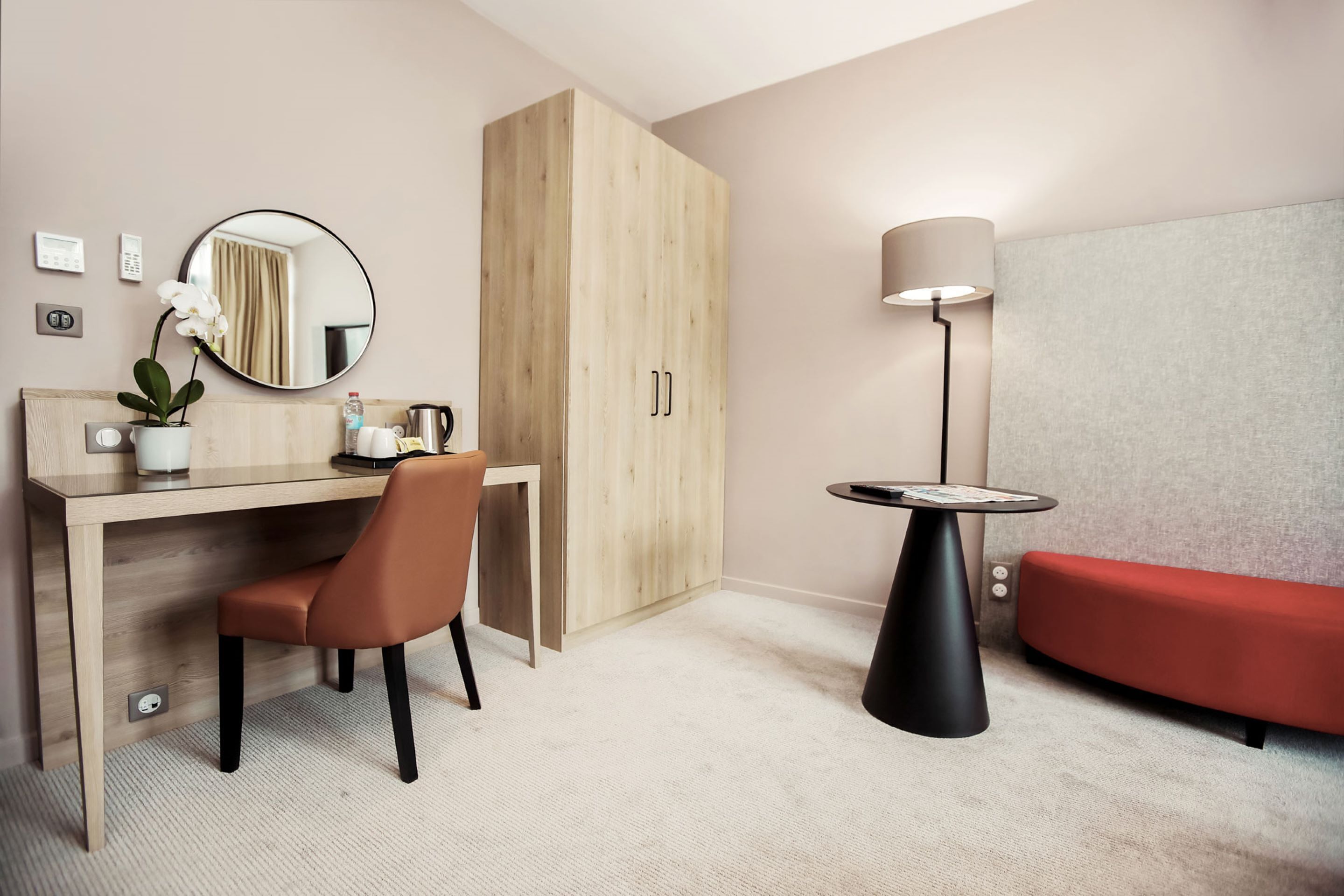Une chambre d’hôtel moderne avec un bureau en bois, une chaise, un canapé rouge, un miroir rond, une lampe et une armoire.