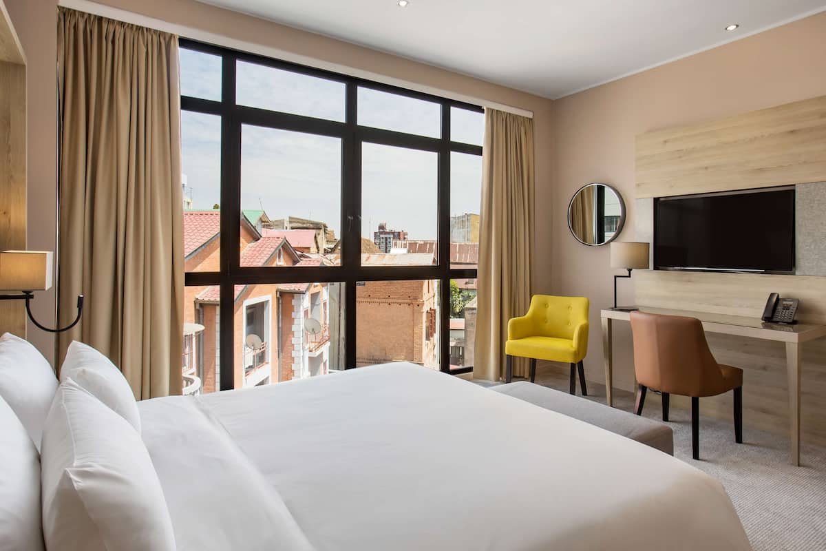 Une chambre d’hôtel avec un grand lit, un bureau avec une chaise, une télévision et une vue sur les immeubles grâce à de grandes fenêtres.
