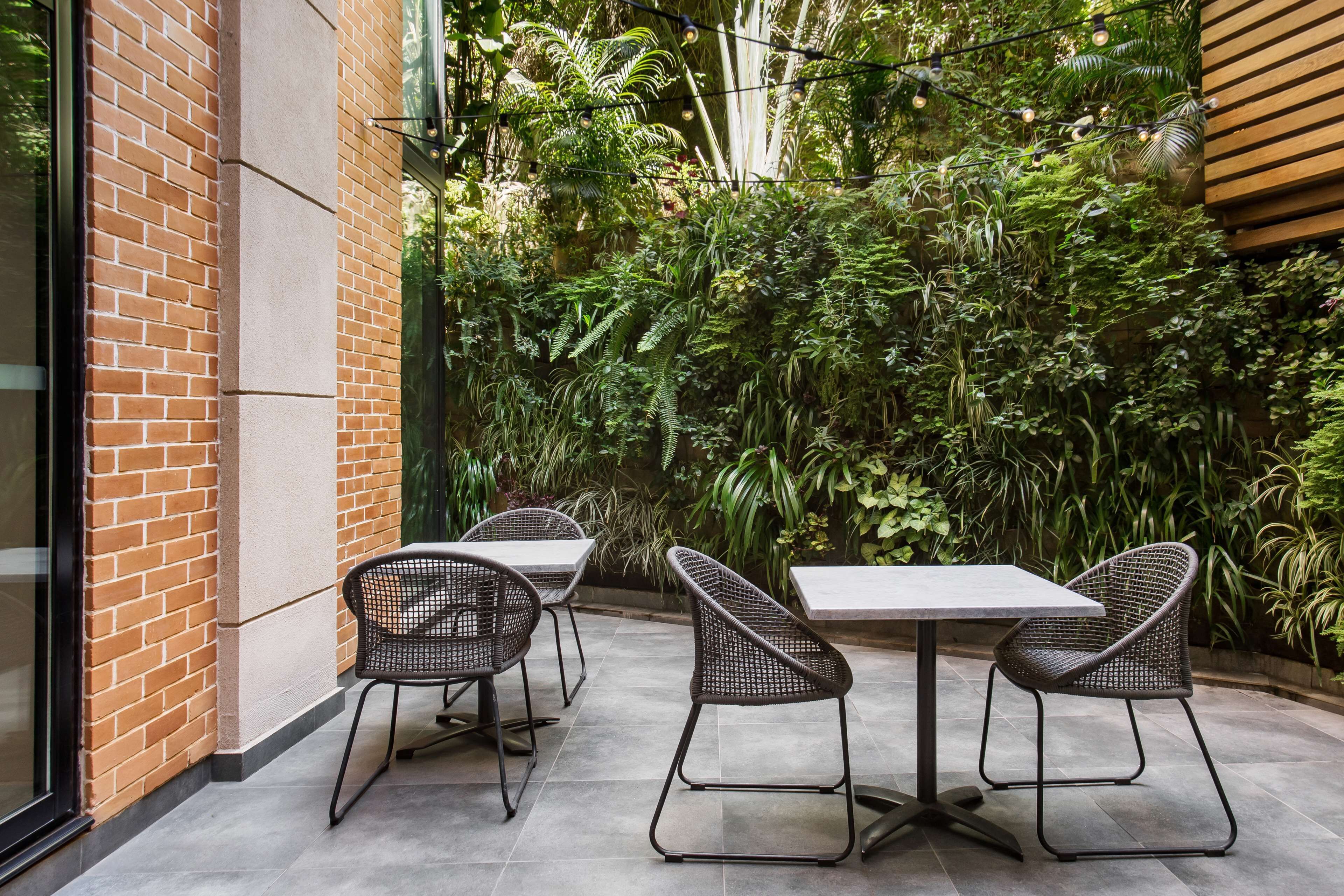 Une terrasse extérieure avec deux tables et quatre chaises, entourée d’une végétation luxuriante et d’un mur de briques.