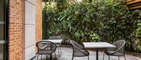 Une terrasse extérieure avec deux tables et quatre chaises, entourée d’une végétation luxuriante et d’un mur de briques.