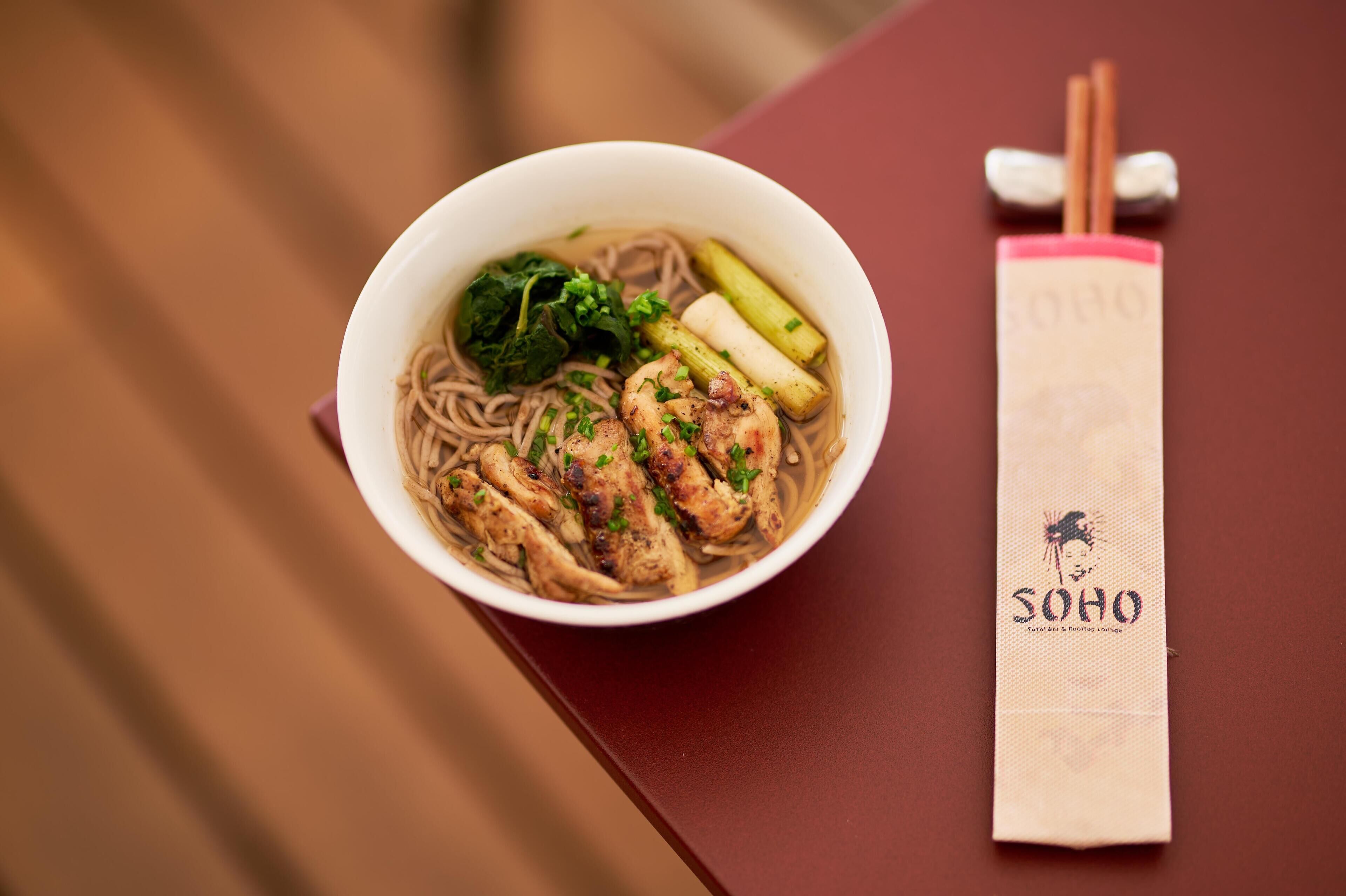 A bowl of noodle soup with vegetables and chicken, accompanied by chopsticks and a napkin with a logo.